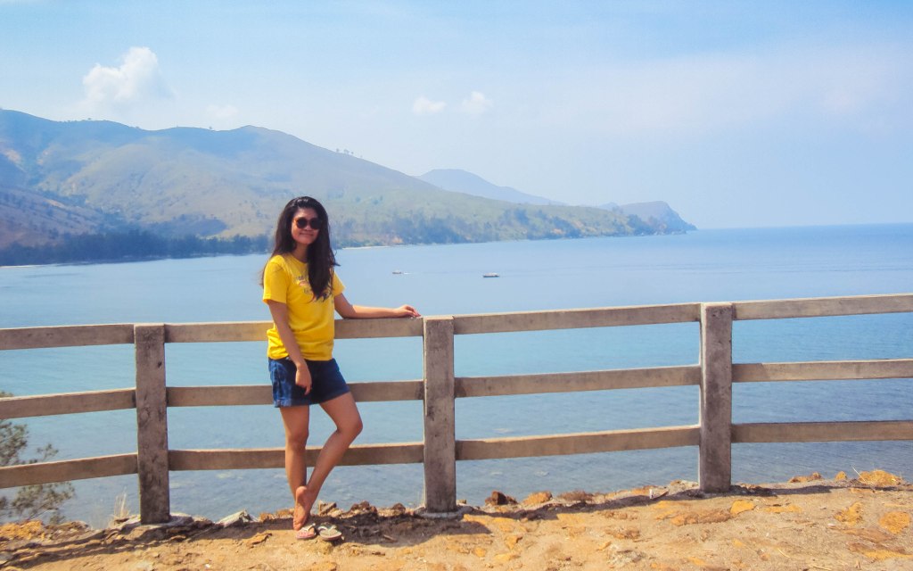 A traveler in a yellow shirt standing by a wooden fence overlooking a serene blue sea and distant mountains on a sunny day.