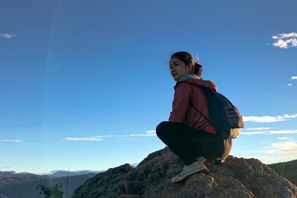 A woman sitting on a rock, overlooking a scenic mountain landscape under a clear blue sky.