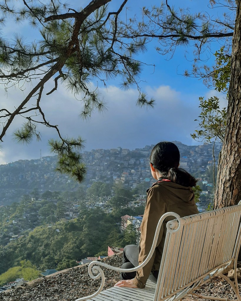 Girl sitting on a bench under a tree looking at an overlooking view of Baguio's hillside