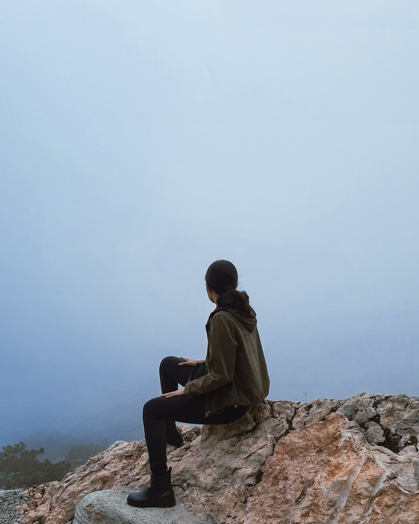 Misty scenery of a girl sitting on a rock and looking at the thick mist in front of her