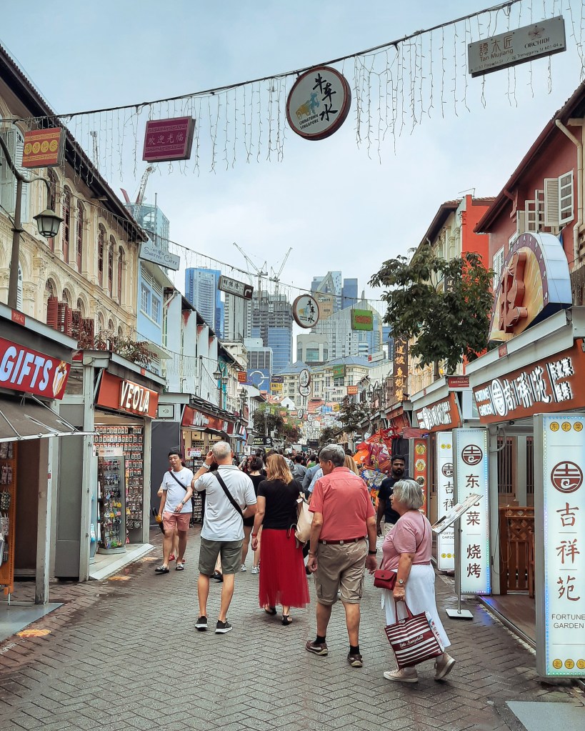 A bustling street in Singapore's Chinatown featuring colorful shops and signboards, with skyscrapers visible in the background. People stroll down the walkway, enjoying the vibrant atmosphere.