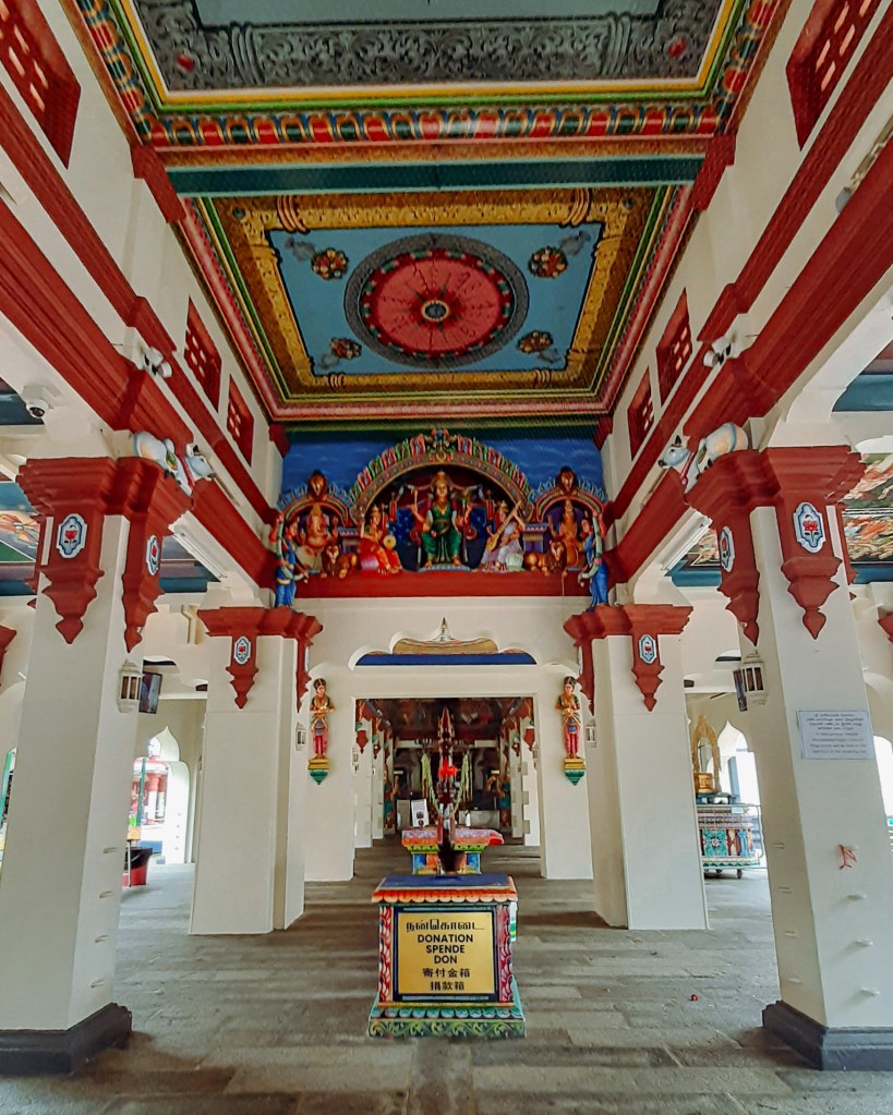 Interior view of a temple featuring colorful painted ceilings and ornate pillars, with a donation box in the center.