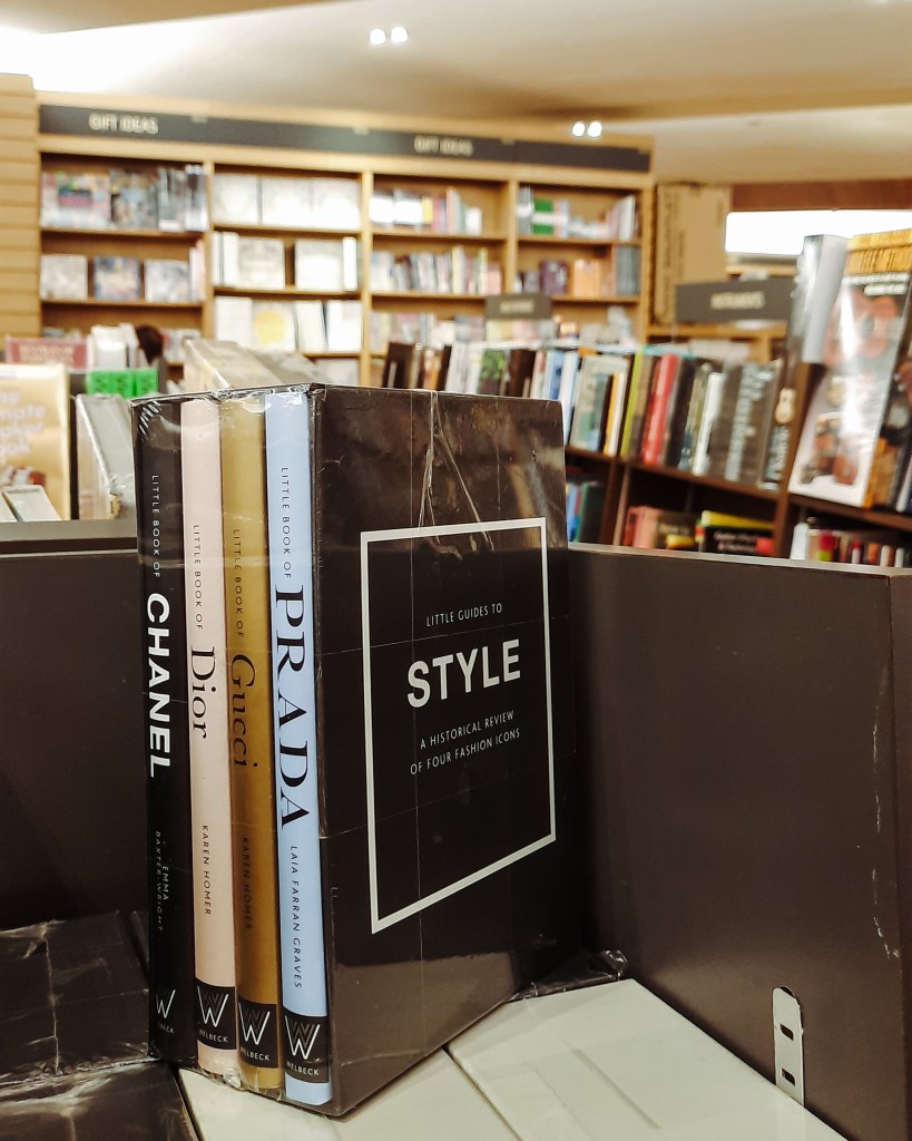 A selection of fashion books on a shelf in a bookstore, including titles on Chanel, Dior, Gucci, and Prada, along with a book titled 'Little Guides to Style: A Historical Review of Four Fashion Icons.'
