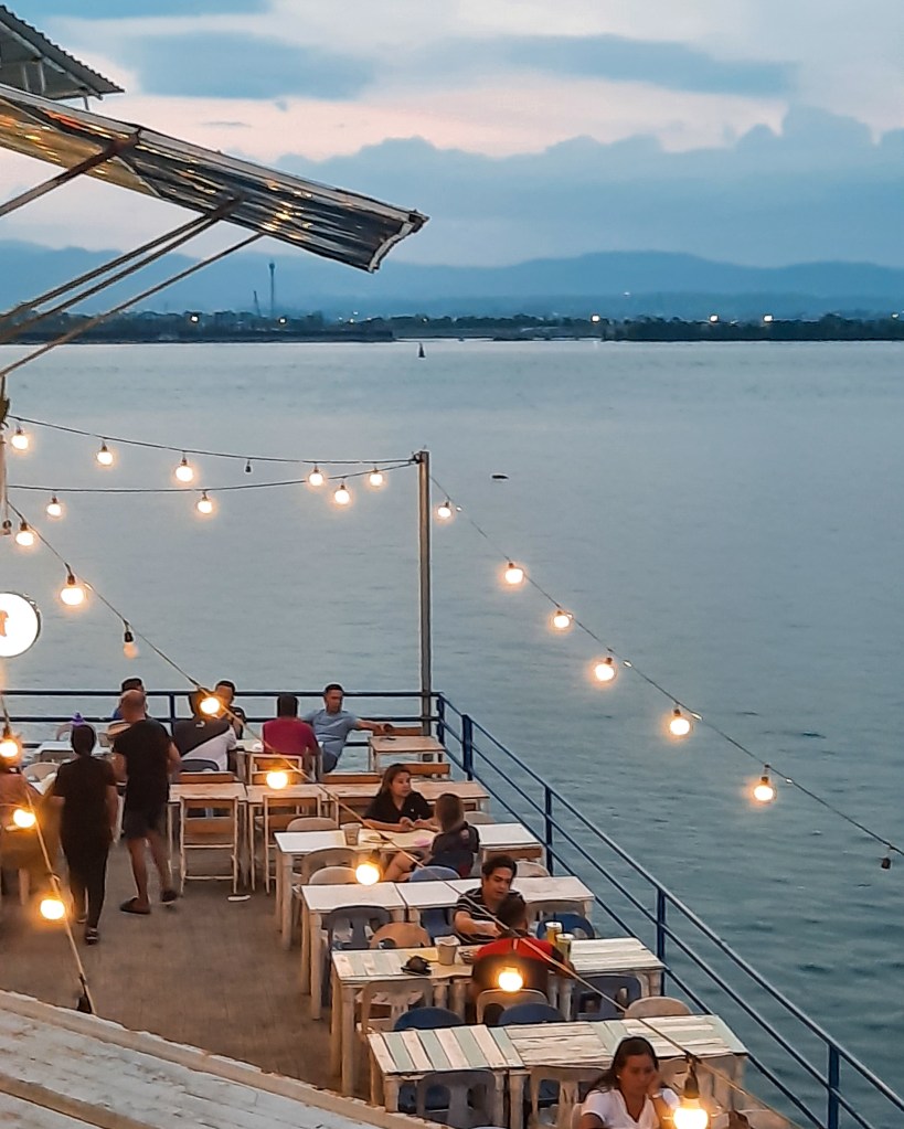 View of a beachfront dining area at sunset, featuring wooden tables and decorative string lights, with patrons enjoying their meals and a scenic waterfront.