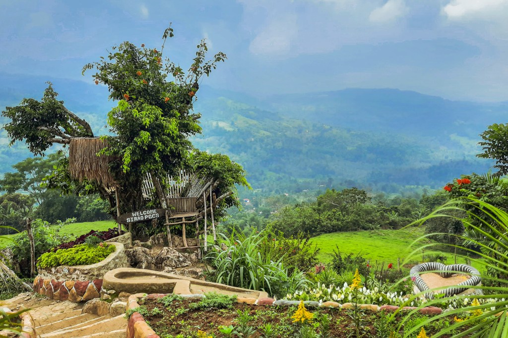 A scenic view of a welcoming sign for a nature destination, featuring a rustic bamboo structure surrounded by lush greenery and mountainous landscape in the background.