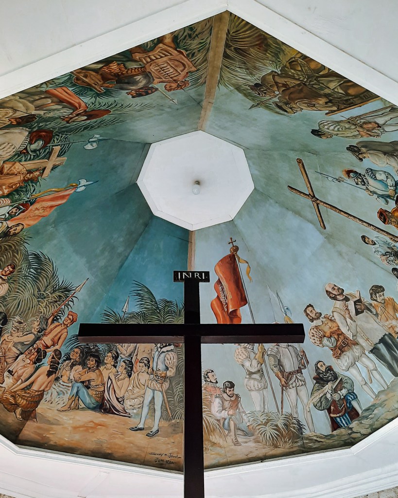 Ceiling mural inside a pavilion featuring a large cross and various historical figures and scenes, depicting the arrival of Ferdinand Magellan in Cebu.