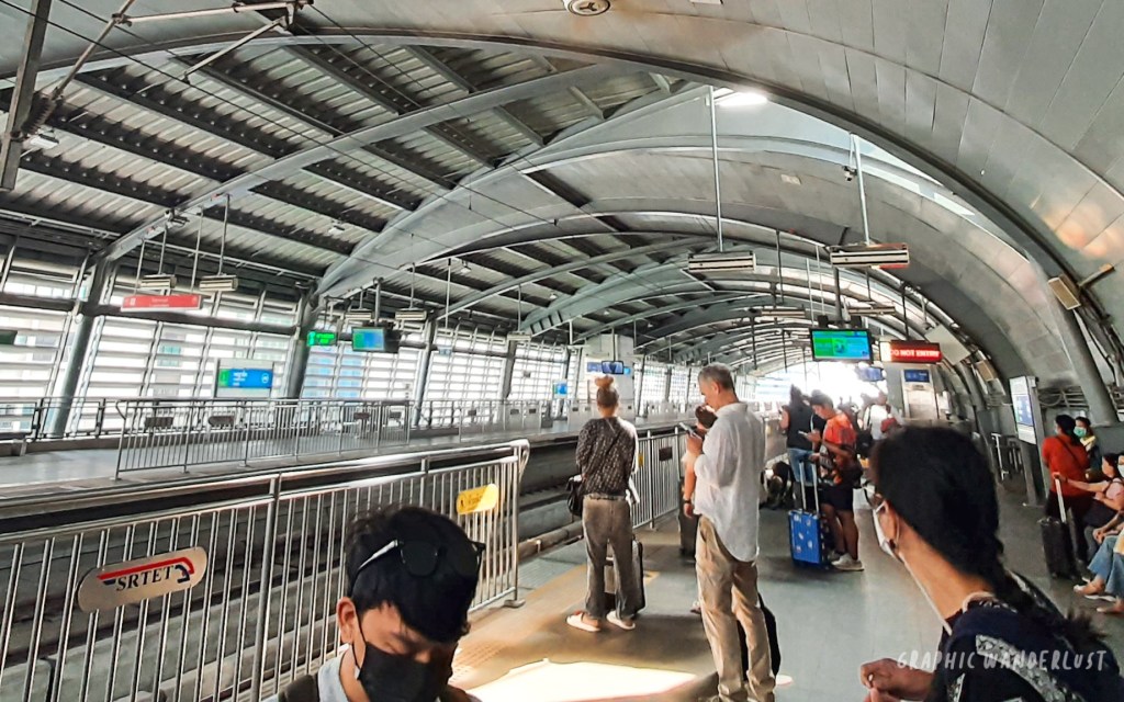 Interior view of a Bangkok Skytrain station with passengers waiting on the platform and electronic information displays visible.