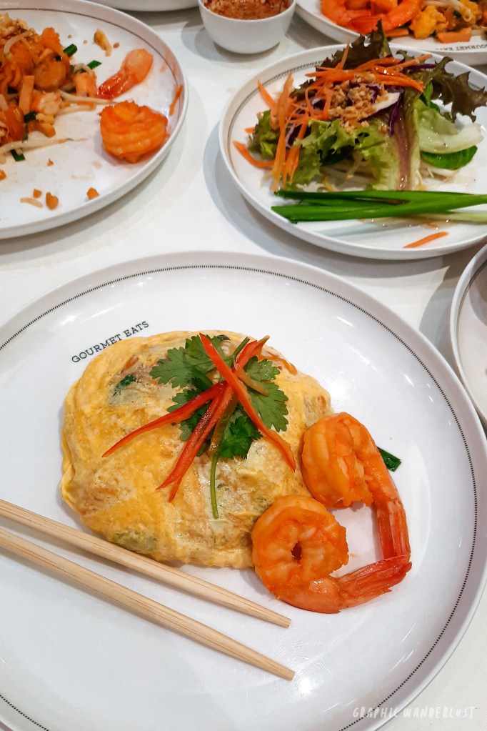 A plate of Pad Thai featuring prawns wrapped in an omelet, garnished with cilantro and red pepper strips, served with chopsticks in a dining setting.