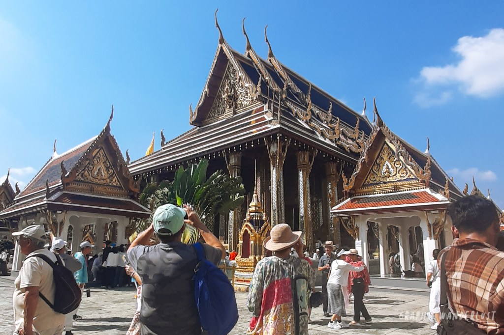 Visitors exploring the ornate architecture of a temple in Bangkok, surrounded by lush greenery and vibrant blue skies.