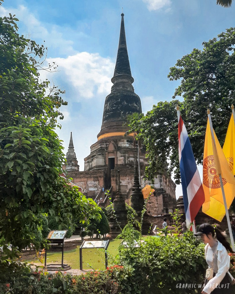 View of a historic temple in Ayutthaya, Thailand, surrounded by greenery and flags, with a clear blue sky in the background.
