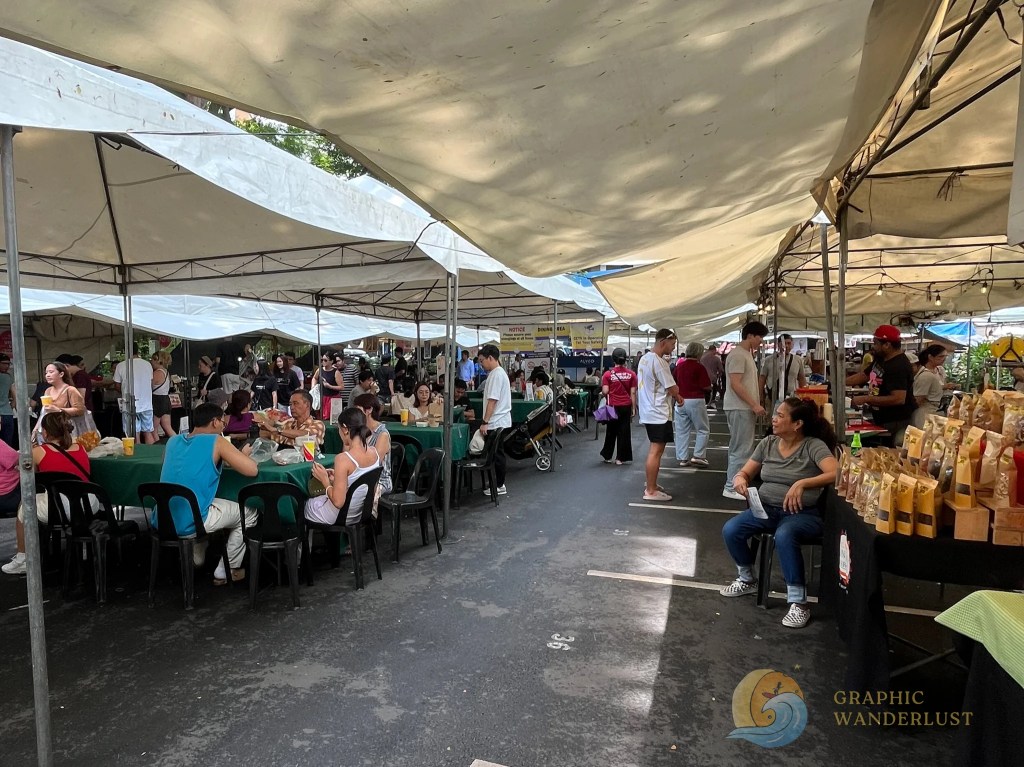 A bustling street market scene in Manila with vendors selling food and products under large tents. People are seated at tables enjoying their meals, while others walk between the stalls.
