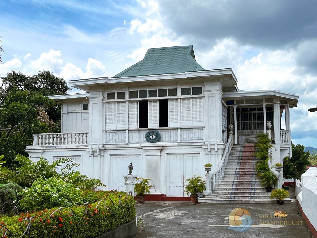 Facade of an ancestral house in Taal, Batangas featuring a white exterior, a green roof, and a staircase surrounded by lush greenery.