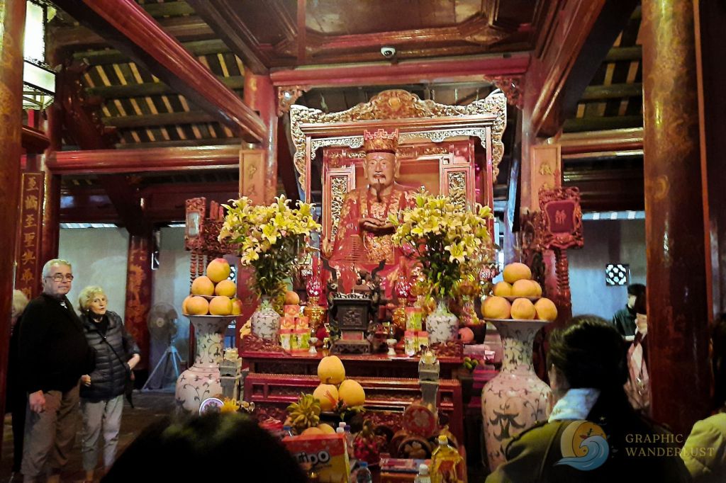 Interior view of a traditional Vietnamese temple, featuring an ornate altar with a statue surrounded by offerings and decorations, and visitors observing.