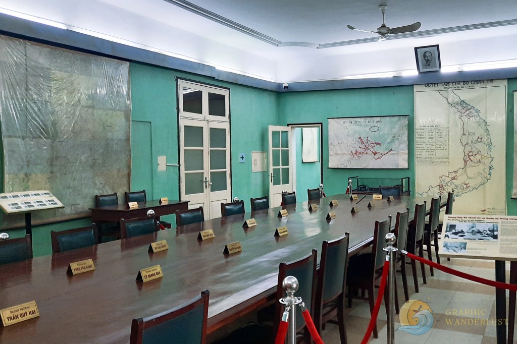 Interior of a historical meeting room at the Imperial Citadel of Thang Long in Hanoi, featuring a large wooden table surrounded by chairs and maps on the walls.