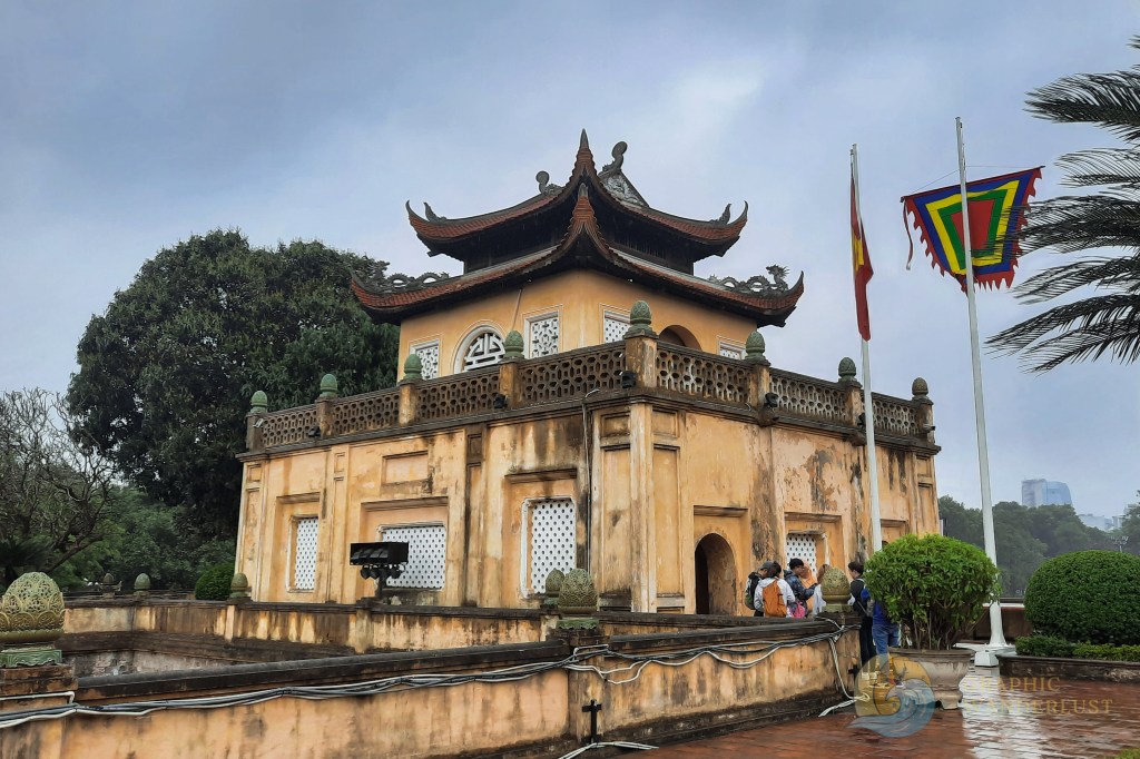 A view of a traditional Vietnamese building surrounded by greenery and flags, with visitors gathered around exploring the area.
