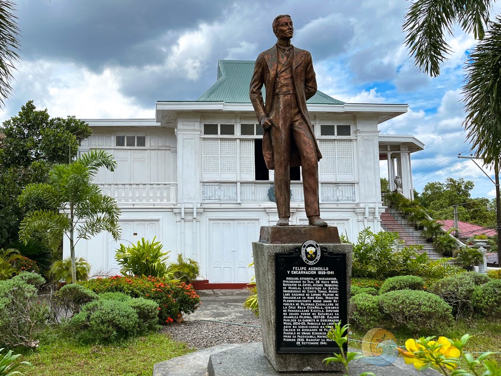 Statue of Felipe Agoncillo, a historical figure, in front of a white Spanish colonial house, surrounded by greenery.