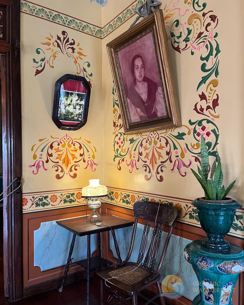 A corner of a historic Spanish colonial house in Taal, Batangas, featuring colorful floral wall paintings, a vintage portrait in a frame, an antique wooden chair, a small table with a lamp, and a decorative plant in a pot.