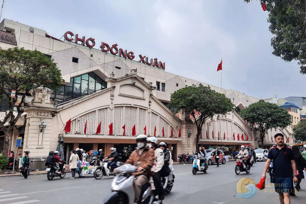 Image of Dong Xuan Market in Hanoi, showcasing the bustling street scene with people and motorbikes in front of the market building with red flags.