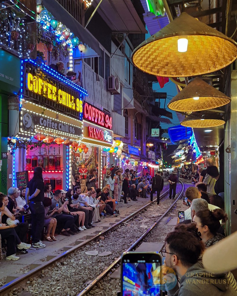 A vibrant street scene in Hanoi's Train Street, features colorful coffee shops and lively crowds gathered along the railway tracks at night.