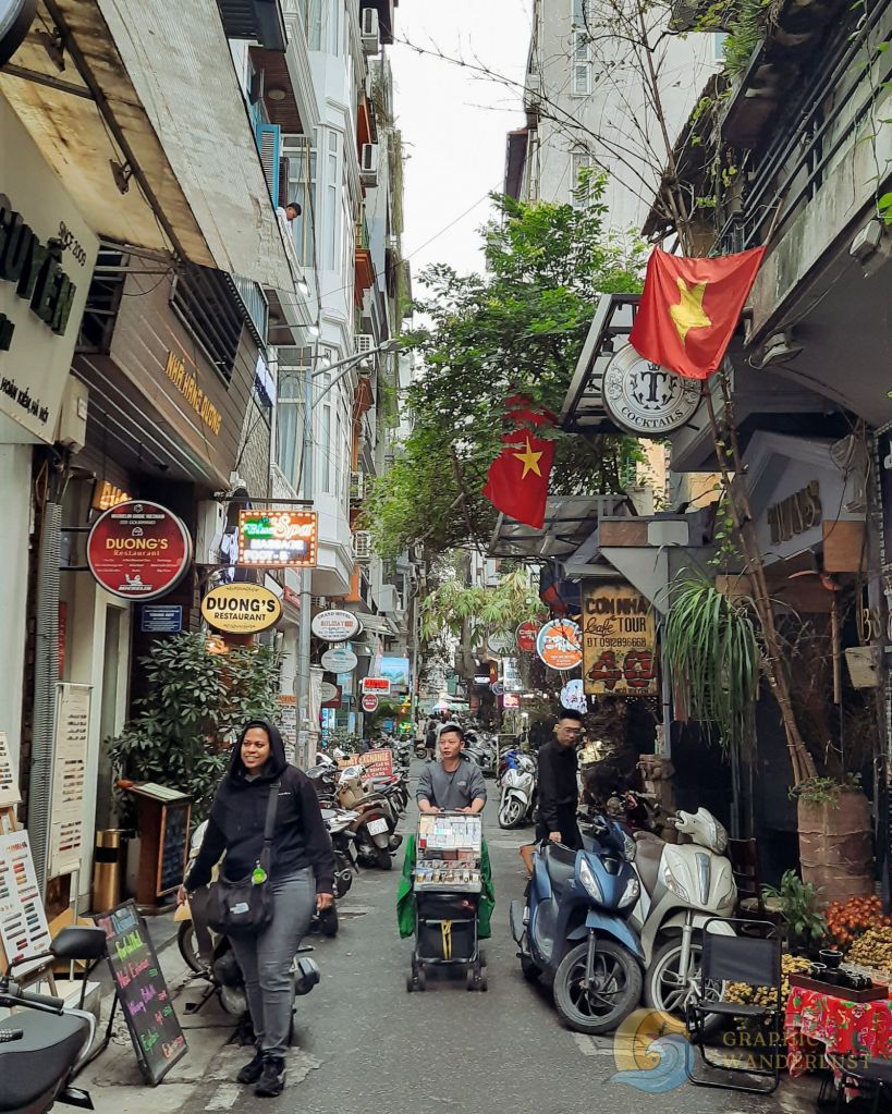 A bustling street in Hanoi's Old Quarter, featuring narrow pathways lined with shops, restaurants, and scooters. People are seen walking and carrying goods, with the Vietnamese flag displayed prominently.
