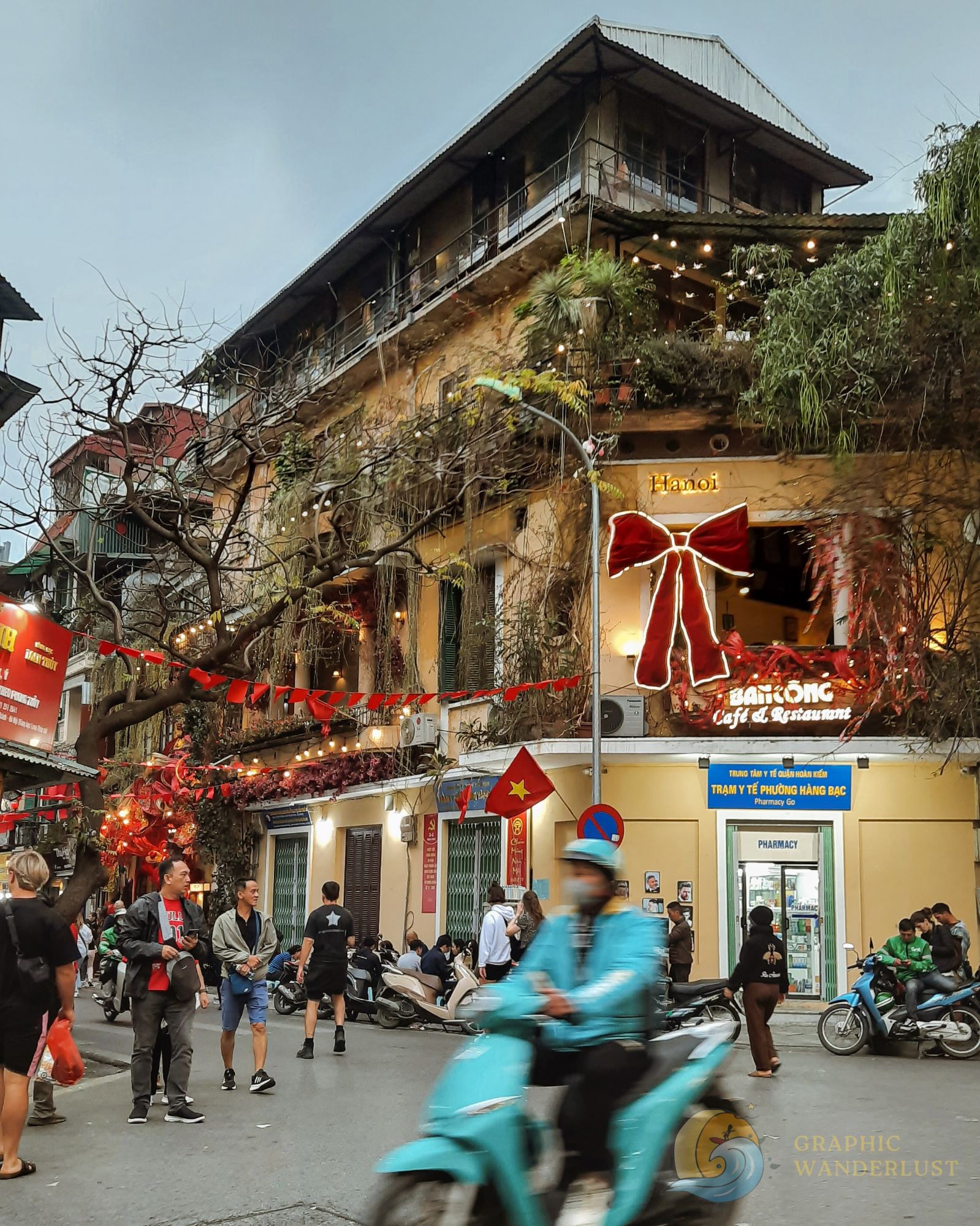 A bustling street scene in Hanoi's Old Quarter, featuring a lively atmosphere with pedestrians, motorcycles, and a charming café adorned with decorations and Vietnamese flags.