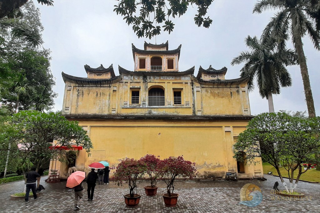 A historical building in Hanoi, Vietnam, showcasing a weathered yellow facade with traditional Asian architectural elements. The structure is surrounded by lush greenery and people walking with umbrellas.