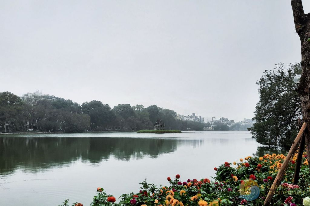 A tranquil view of Hoan Kiem Lake in Hanoi, Vietnam, framed by colorful flowers in the foreground and a cloudy sky above.