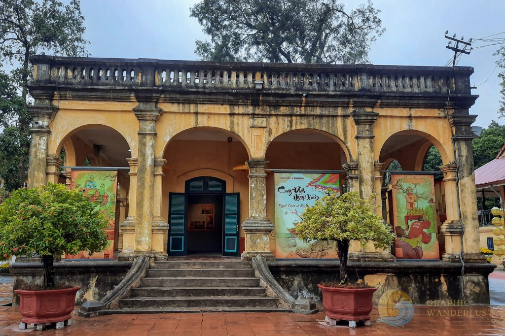 Exterior view of a historic building with yellow walls and green shutters, featuring an entrance with stone steps and decorative plants in pots.