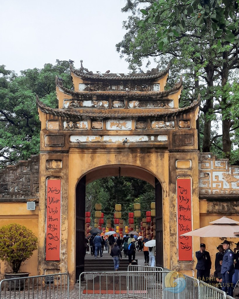 A historic entrance gate to a cultural site in Hanoi, Vietnam, featuring traditional architectural elements and inscriptions, with visitors entering under lanterns amidst lush greenery.