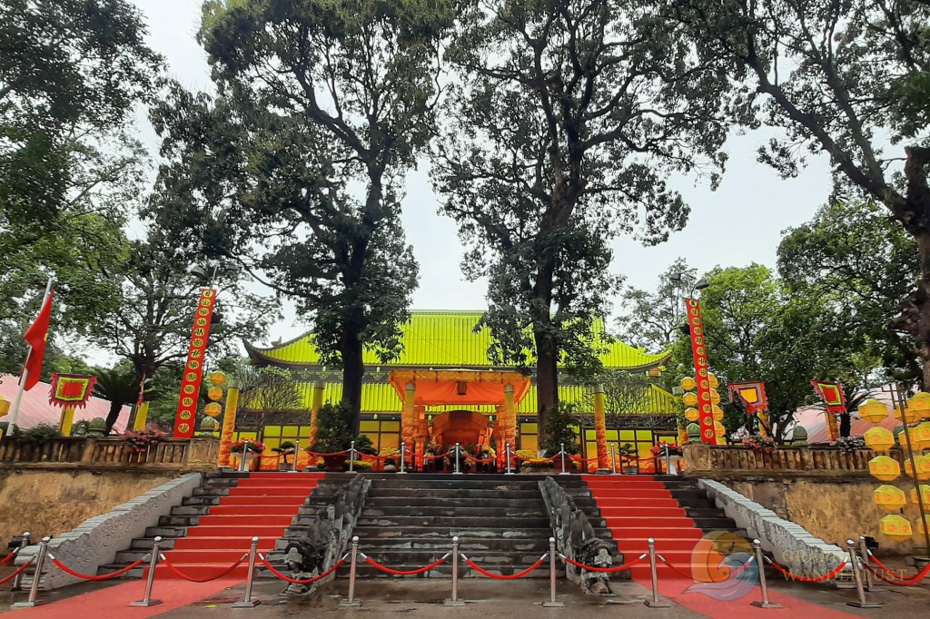 Image of a temple entrance in Vietnam, featuring ornate decorations, colorful banners, and lush trees surrounding the area.