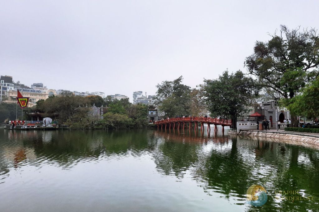 A scenic view of Hoan Kiem Lake in Hanoi, featuring a red wooden bridge leading to a small island with a temple, surrounded by lush greenery and urban buildings in the background.