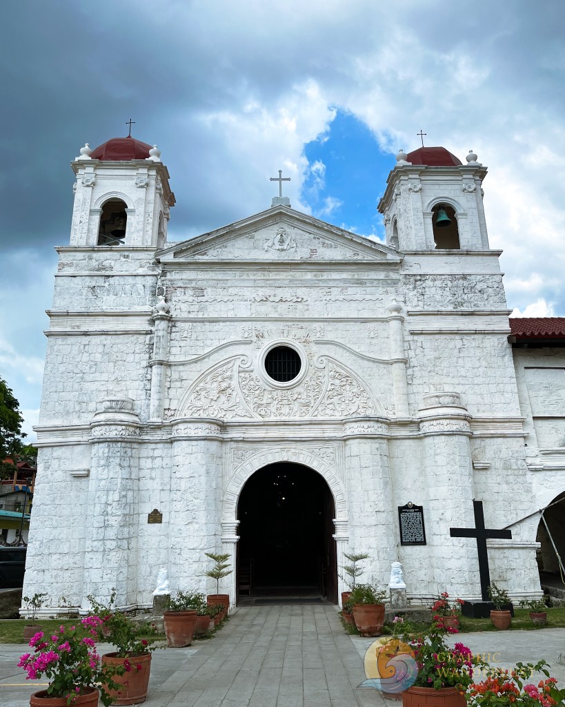Facade of the Archdiocesan Shrine of Our Lady of Caysasay, showcasing its white stone exterior, baroque details, and red domes under a cloudy sky.