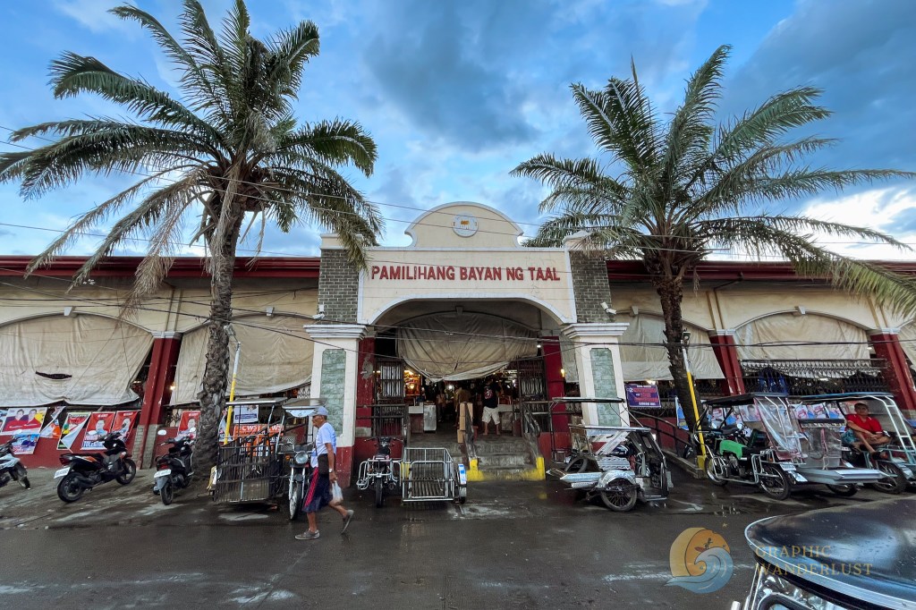 Entrance to the Taal Public Market with palm trees and tricycles parked outside.