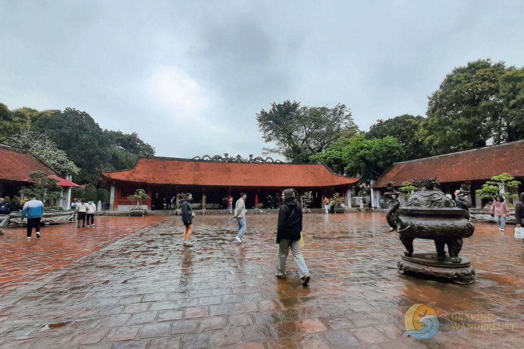 A courtyard view of the Temple of Literature in Hanoi, Vietnam, featuring visitors walking on a rainy day with traditional architecture in the background.