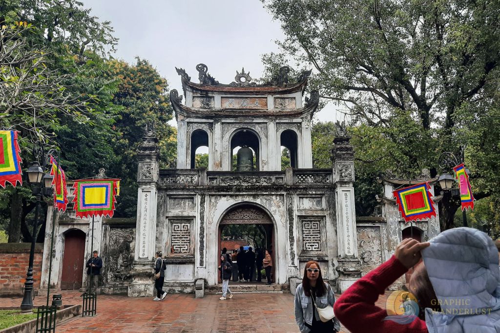 Entrance gate of the Temple of Literature in Hanoi, Vietnam, decorated with colorful flags and surrounded by trees and visitors.
