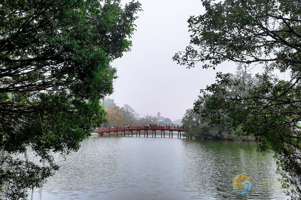 View of Hoan Kiem Lake framed by lush greenery, featuring the red Huc Bridge in the distance.