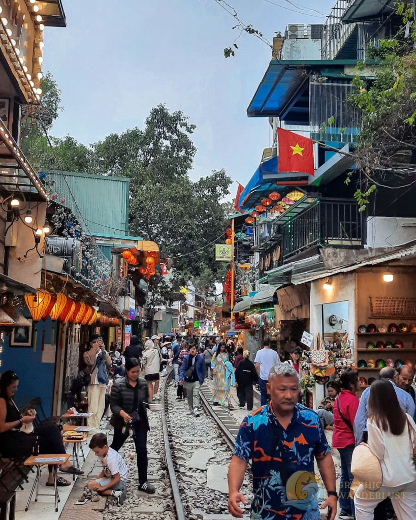 A bustling scene at Train Street in Hanoi, Vietnam, filled with locals and tourists exploring cafes and shops along the railway tracks.