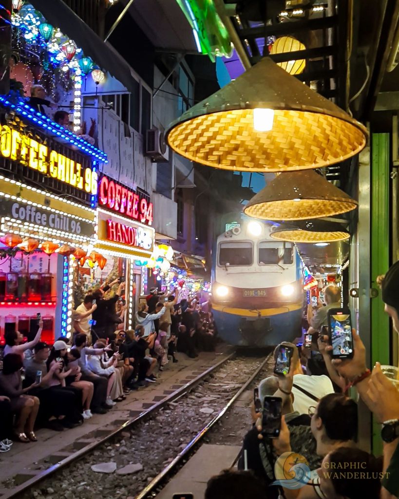 A train passing through Hanoi's Train Street, surrounded by colorful cafés and enthusiastic spectators capturing the moment on their phones.