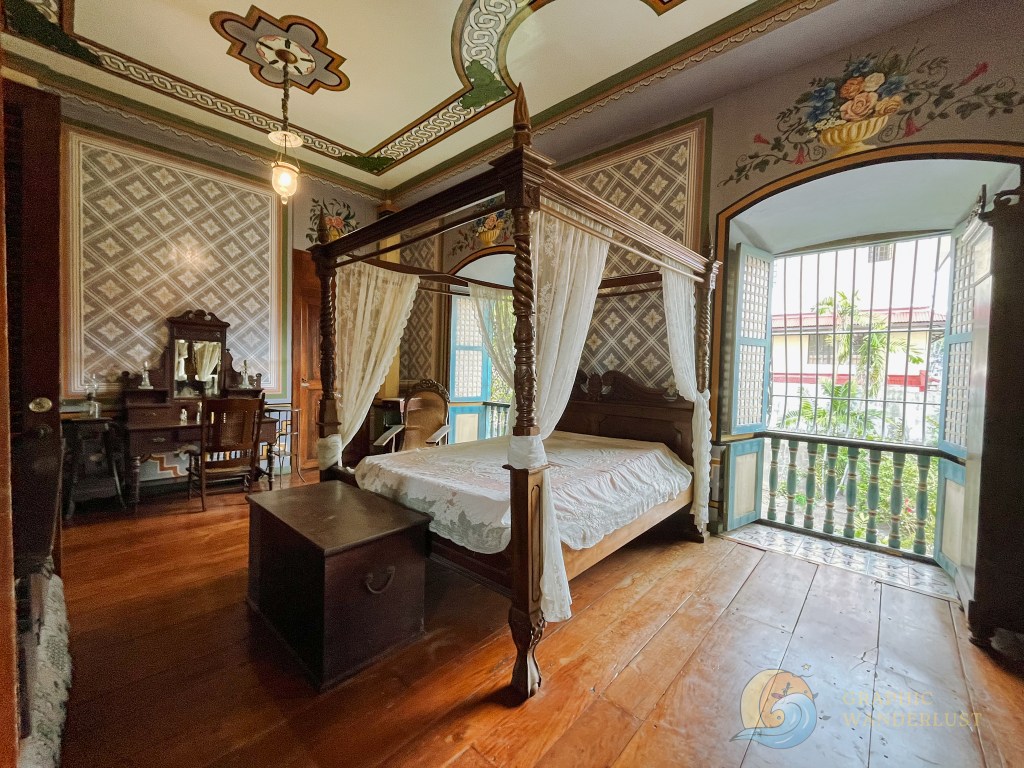 Interior view of a historical bedroom featuring a four-poster bed with white drapes, vintage wooden furniture including a dresser and a chair, and colorful patterned walls with floral designs.
