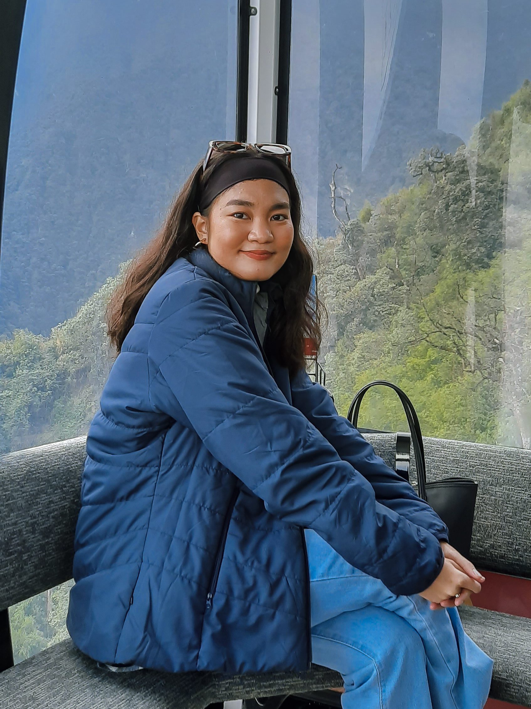 A woman with long hair wearing a blue jacket and headband, sitting in a gondola with a scenic mountain view in the background.