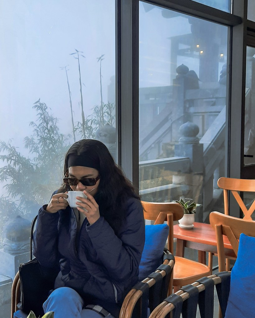 A person sitting indoors, wearing sunglasses and a dark jacket, holds a cup while looking down, with a misty landscape visible through the window behind.
