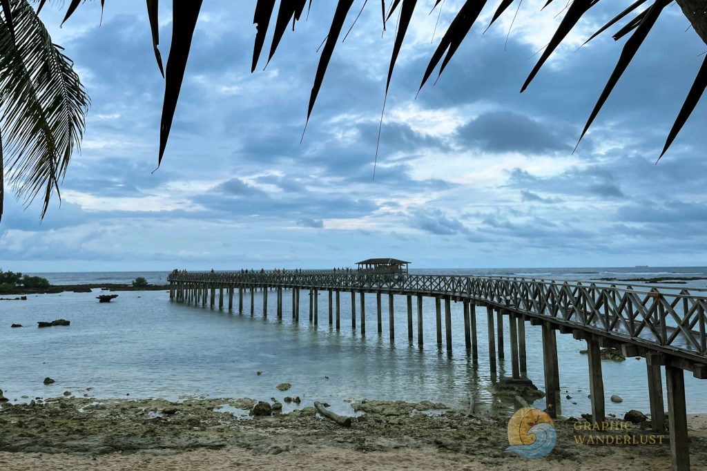 Scenic view of a wooden pier extending into calm waters under a cloudy sky, surrounded by lush greenery.