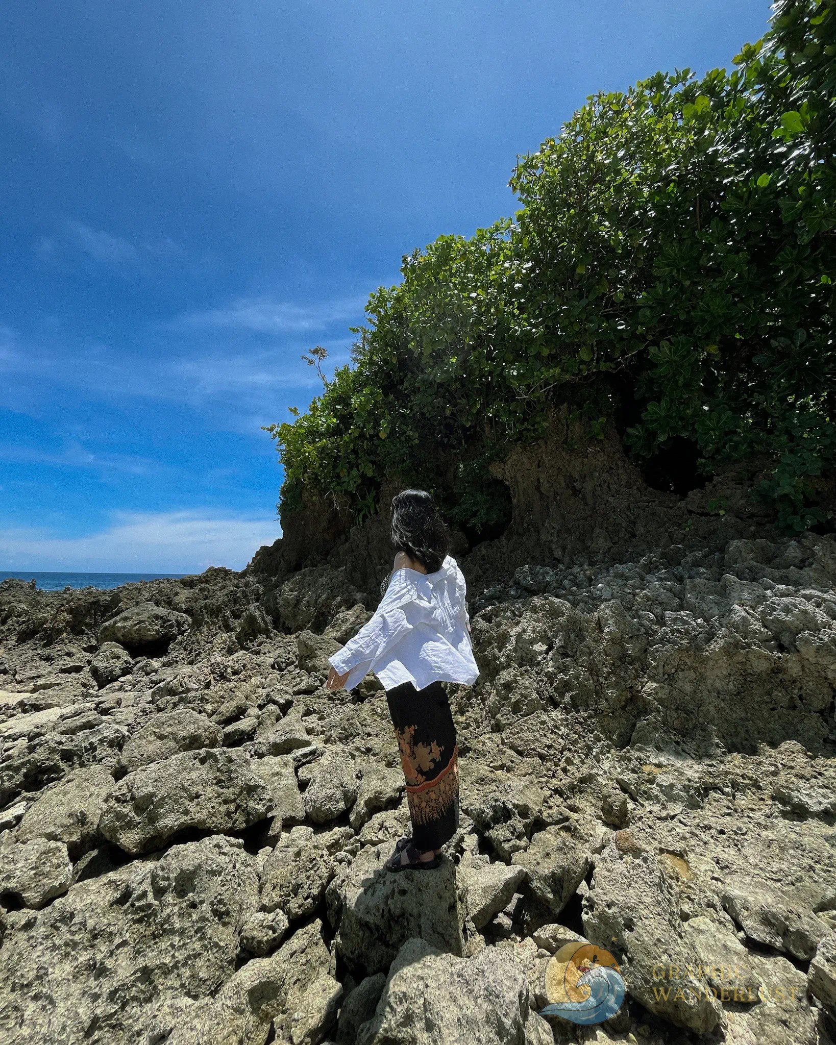 A woman standing atop of rock formations in a shore with background of lush greenery and clear blue sky and blue waters in the distance.