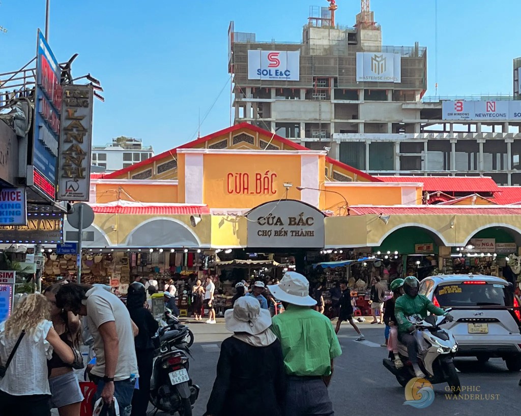 A bustling street market scene featuring the entrance to a market labeled 'CỬA BẮC'. Numerous shoppers and vendors are present, with scooters and motorcycles visible in the foreground. In the background, modern buildings are under construction.