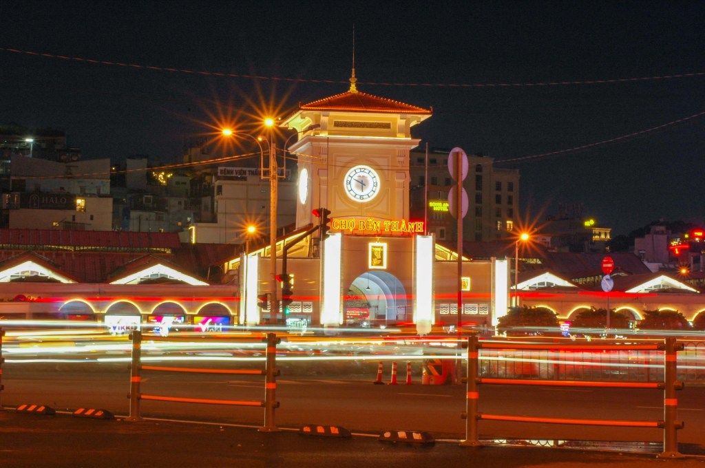 A brightly lit night view of Ben Thanh Market in Ho Chi Minh City, featuring its iconic clock tower and illuminated architecture, with light trails from passing vehicles in the foreground.