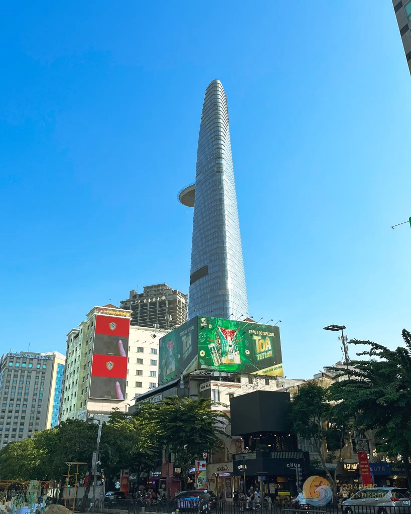 View of a tall skyscraper against a clear blue sky, surrounded by modern buildings and vibrant advertisements.