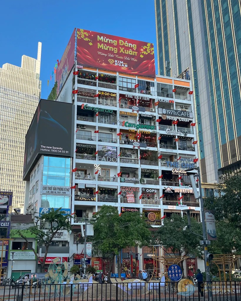 A colorful multi-story building adorned with various signs for cafes and shops, featuring decorations for a celebration. The top banner reads 'Mừng Đăng Mừng Xuân'.