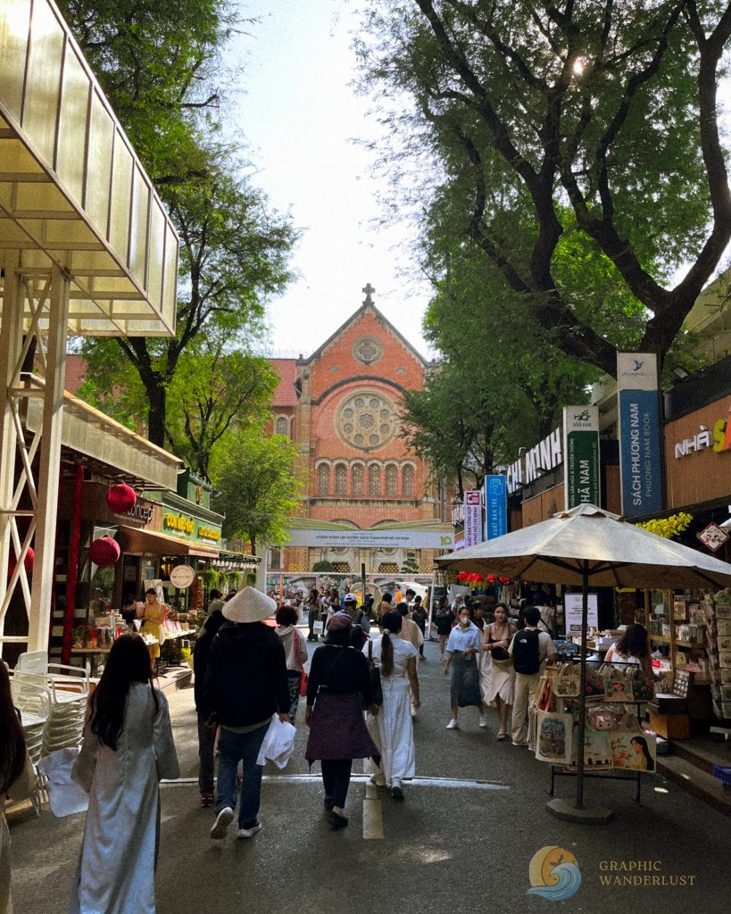 A bustling street scene with people walking between shops and stalls, featuring a large historic building in the background, surrounded by lush trees.
