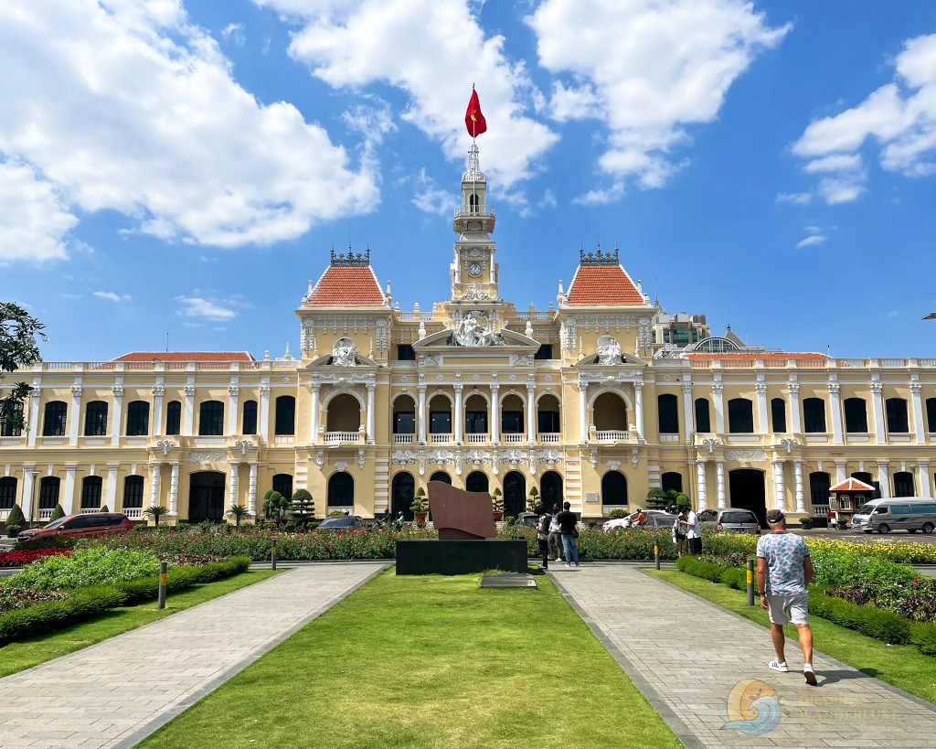 Historic building with French colonial architecture, featuring a tall clock tower, a flag on top, and surrounded by well-maintained gardens and pathways.