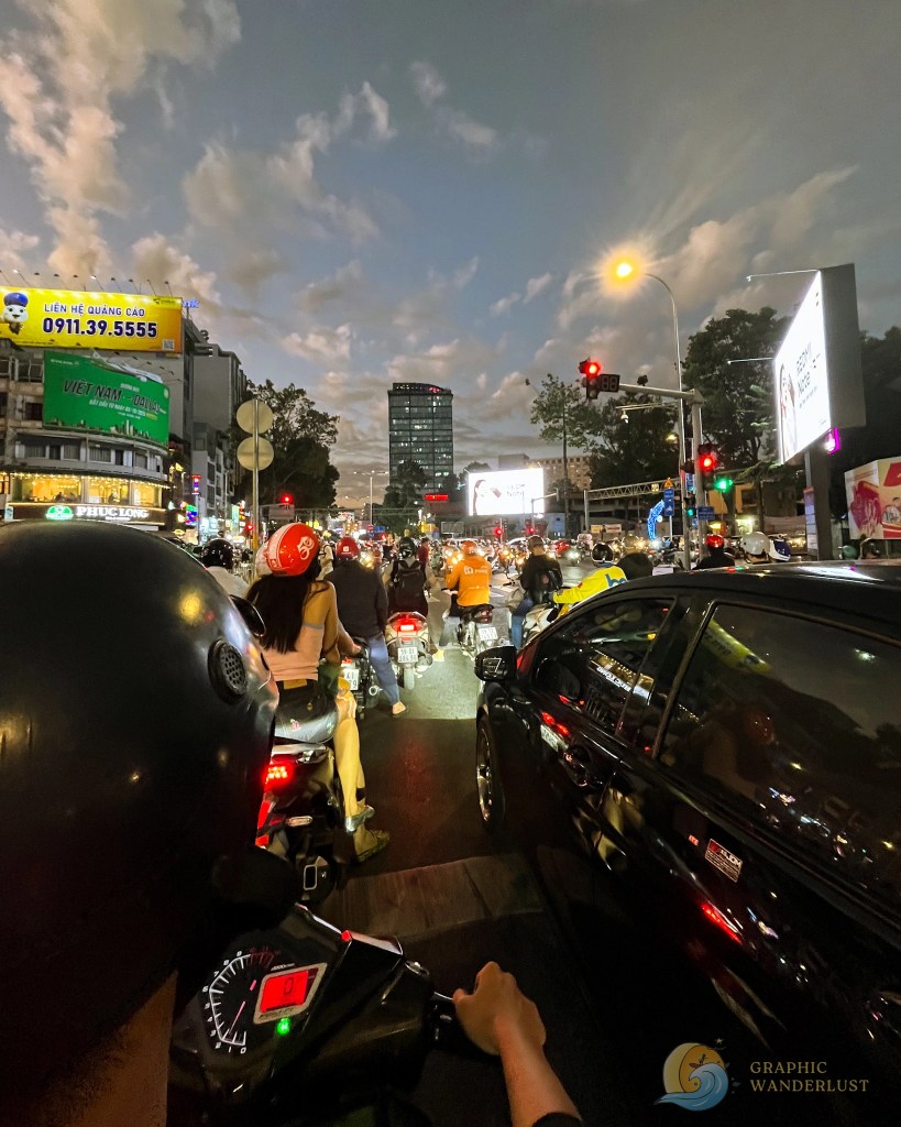 A busy street in a city during evening rush hour, filled with motorcycles and cars stopped at a traffic light, with billboards and a cloudy sky in the background.